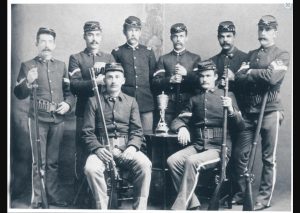 A black and white photo of the Fort Mackinac rifle team from 1886. The soldiers are posed for the photo, holding rifles, with a trophy between them.