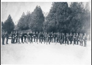 A black and white photograph of 1880s soldiers standing in a line with muskets and rifles in front of them. 